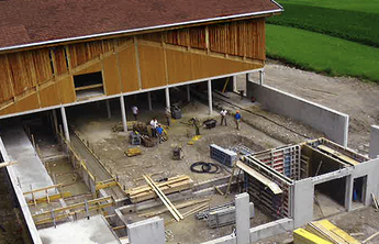 Eine Baustelle mit einem Gebäude im Rohbau, das eine Holzverkleidung hat. Arbeiter sind an verschiedenen Stellen aktiv, während Baumaterialien wie Bretter und Betonumschließungen verwendet werden. Im Hintergrund sind grüne Felder sichtbar.