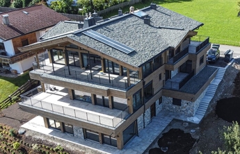 Ein modernes, mehrstöckiges Gebäude mit Holz- und Steinverkleidung ist zu sehen. Es besitzt große Balkone mit Geländer und ein steiles Dach, das mit Schindeln bedeckt ist. Der Gartenbereich ist noch nicht vollständig angelegt, und im Hintergrund ist eine grüne Wiese sichtbar.