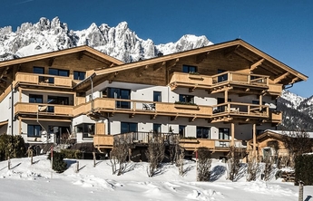 Ein dreigeschossiges Chalet im alpinen Stil mit Holzbalustraden und mehreren Balkonen, umgeben von schneebedeckten Landschaften. Im Hintergrund erheben sich beeindruckende Berge unter einem blauen Himmel. Das Gebäude strahlt Gemütlichkeit und alpine Tradition aus.
