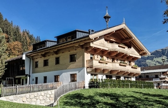 Ein traditionelles alpenländisches Haus mit Holzbalustraden und einem charakteristischen Dach. Die Fassade ist aus hellem Holz und ergänzt durch große Fenster. Umgeben von Wiese und Bäumen, vermittelt es ein einladendes und gemütliches Ambiente.