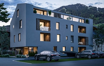 Moderne, mehrstöckige Wohngebäude in einem ländlichen Alpen-Setting. Die Fassade ist grau mit Fenstern, die Licht ins Innere lassen. Vor dem Gebäude stehen mehrere Autos auf einem Parkplatz. Im Hintergrund sind sanfte Hügel und Bäume sichtbar.