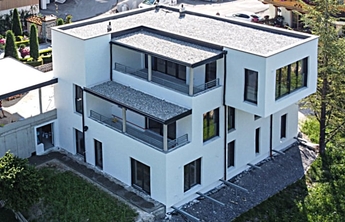 Moderne, dreistöckige Wohngebäude in weißer Fassade, mit großen Fenstern und Balkonen. Das Dach ist mit Kies bedeckt. Umgeben von einem grünen Garten und angrenzenden Straßen.