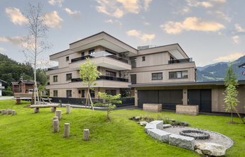 Ein modernes, mehrstöckiges Gebäude aus Holz, umgeben von Grünflächen und Bäumen. Im Vordergrund sind verschiedene Sitzgelegenheiten und Steinelemente sichtbar. Die Architektur ist klar und angenehm, mit Balkonen, die eine Aussicht auf die Umgebung bieten.